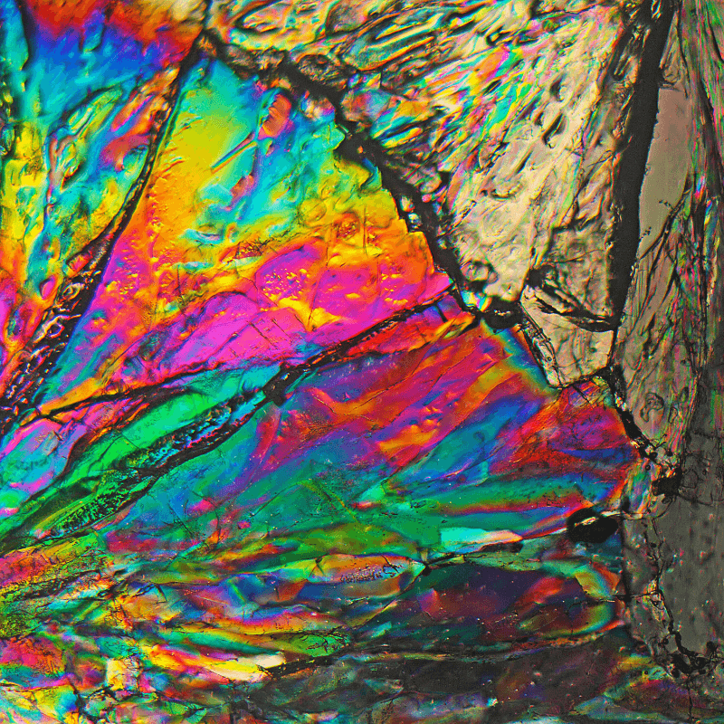 Mikroskopische Aufnahme einer kristallinen Struktur unter polarisiertem Licht mit intensiv leuchtenden Regenbogenfarben wie Pink, Gelb, Grün und Blau auf teils dunklem, strukturiertem Untergrund.